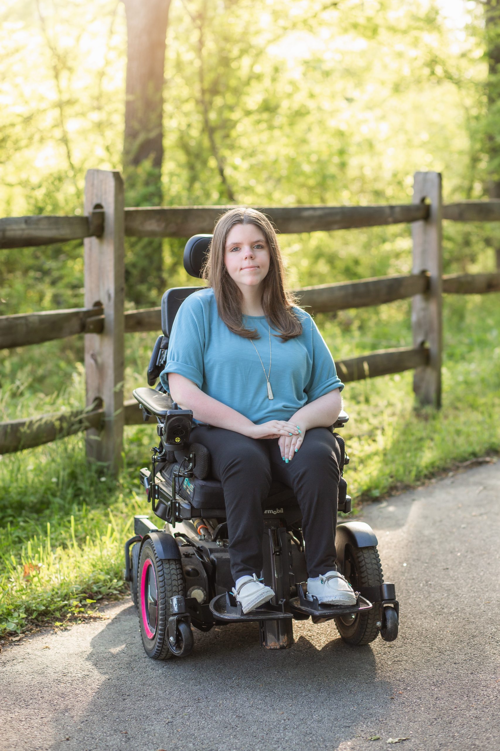 Haley Hayes, TWYPP Session 16 co-moderator, seated in a power wheelchair on an outdoor path. She has long brown hair and is wearing a teal top, black pants, and a long pendant necklace. A wooden fence and sunlit trees are visible in the background.