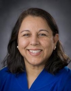 Headshot of Dr. Deeksha Bali, smiling, wearing a royal blue top, with dark hair worn down, against a gray studio background.