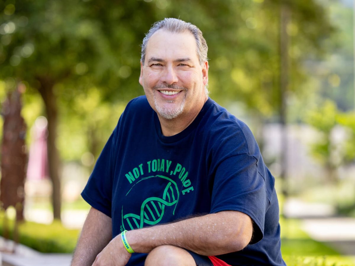 Dwayne Wilson smiling outdoors, seated with arms resting on his knee, wearing a navy blue t-shirt printed with 'Not Today, Pompe' and a green DNA helix graphic, and a green awareness wristband. Trees and a park path are blurred in the background.