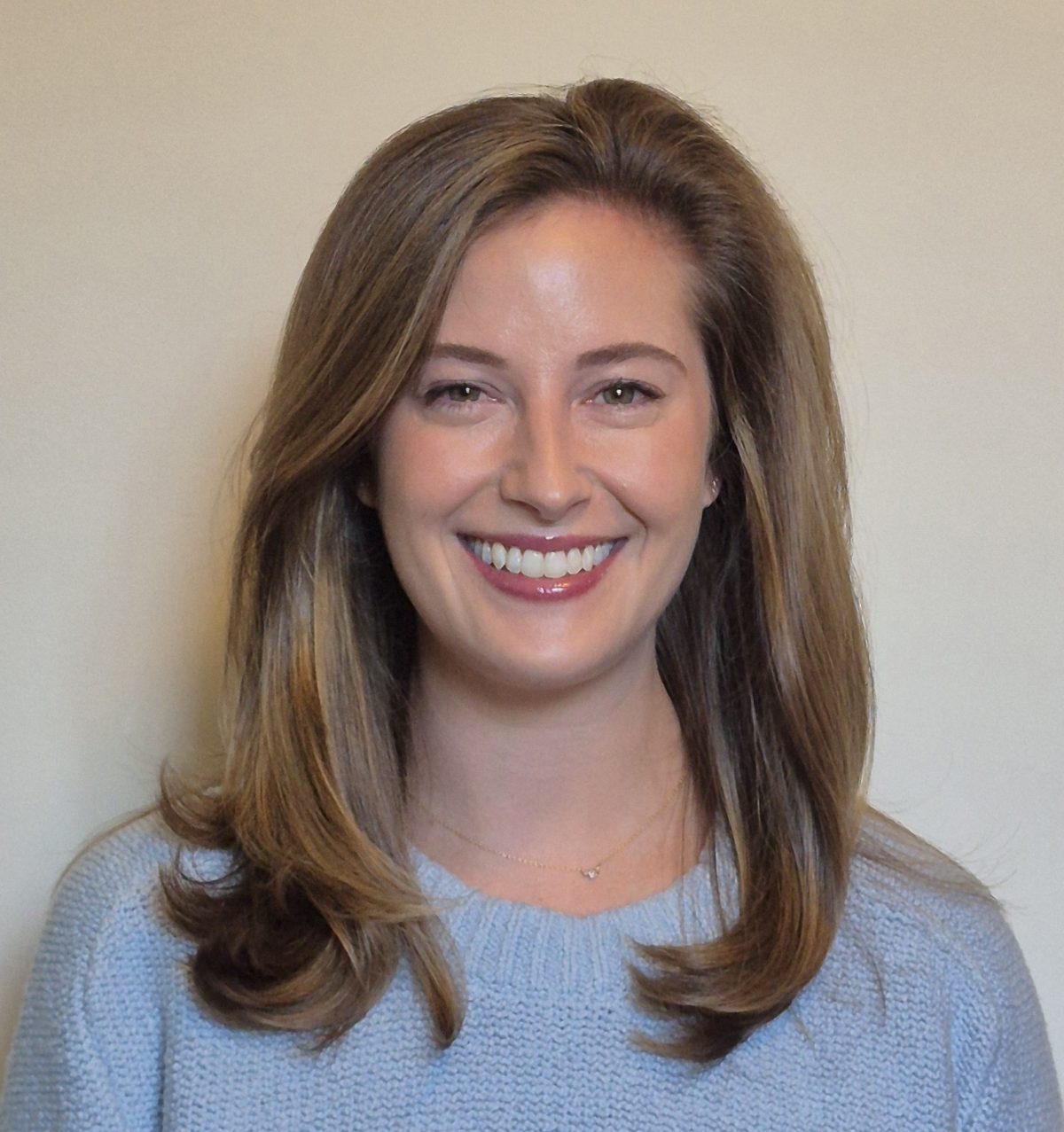 Headshot of Kristen Haggarty-Waite, smiling, with shoulder-length brown hair, wearing a light blue sweater, against a plain neutral background.