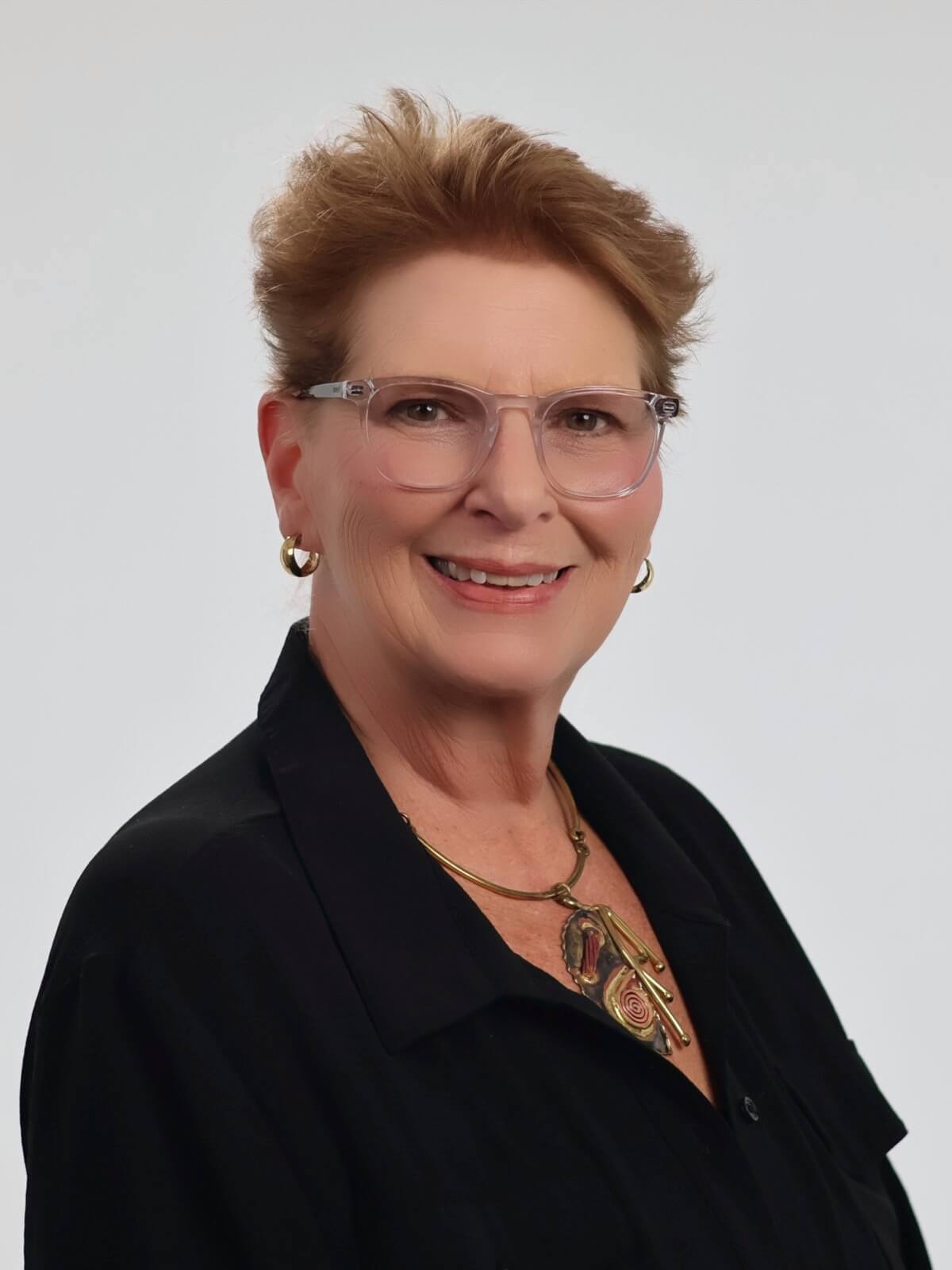 Marsha Zimmerman smiling in a headshot, wearing glasses and a black top with a statement necklace, against a light gray background.