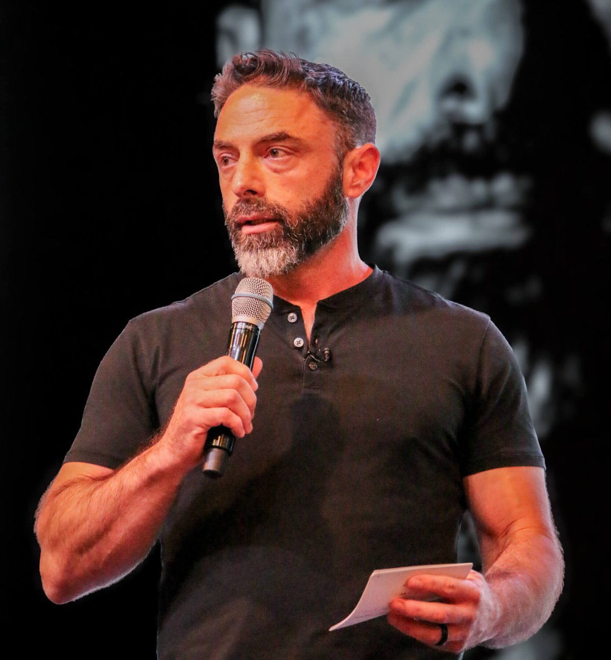 Brandon_Mancine Brandon Mancine speaking into a handheld microphone on stage, wearing a black short-sleeved henley and holding a small note card.