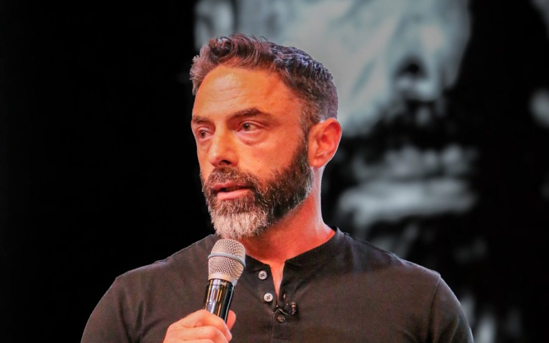 Brandon Mancine speaking into a handheld microphone on stage, wearing a black short-sleeved henley and holding a small note card, with a large black-and-white portrait projected on the screen behind him.